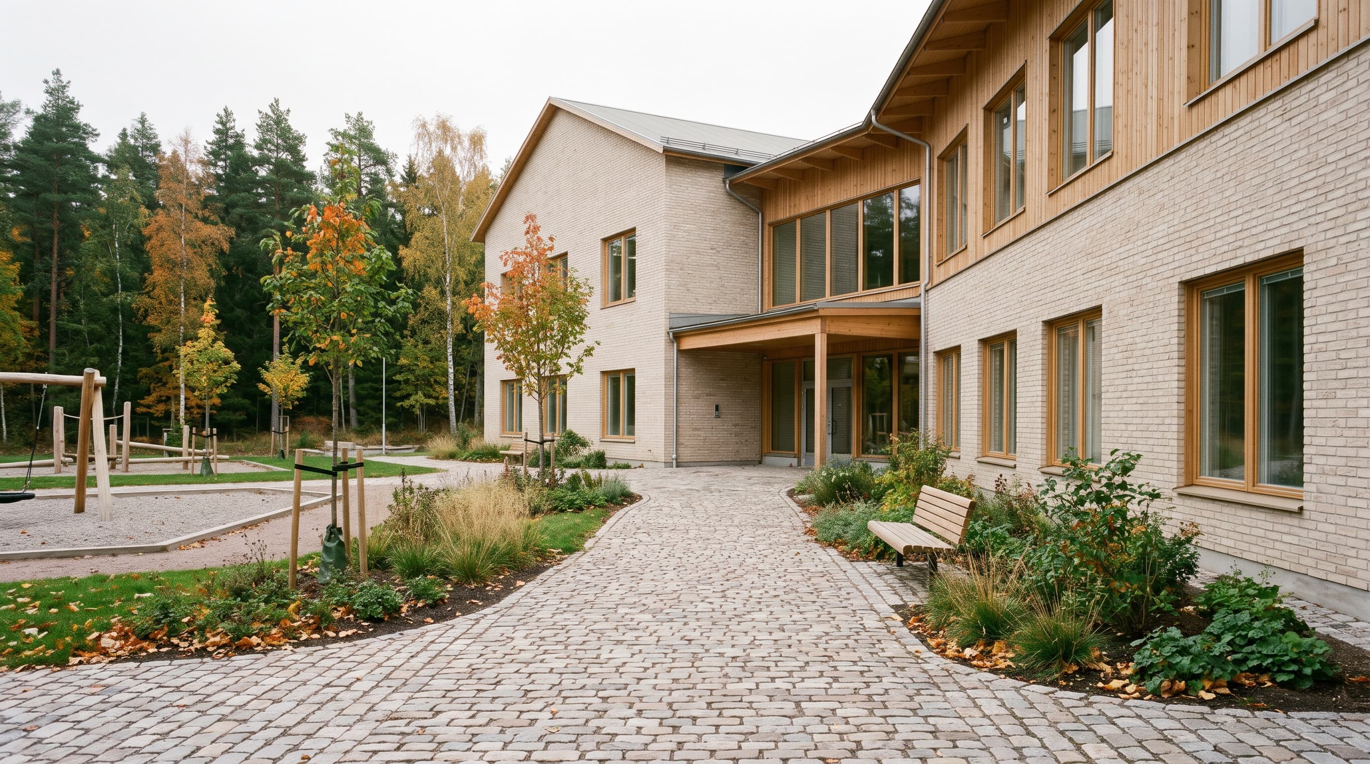 Nybyggd skola i ljust tegel och trä med kullerstensgång, bänk, planteringar och lekplats mot höstskog – en färdigställd skolbyggnad